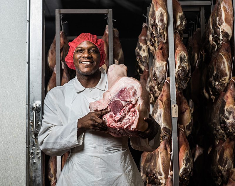 Volpi employee holding a cured ham with more hams hanging in the background