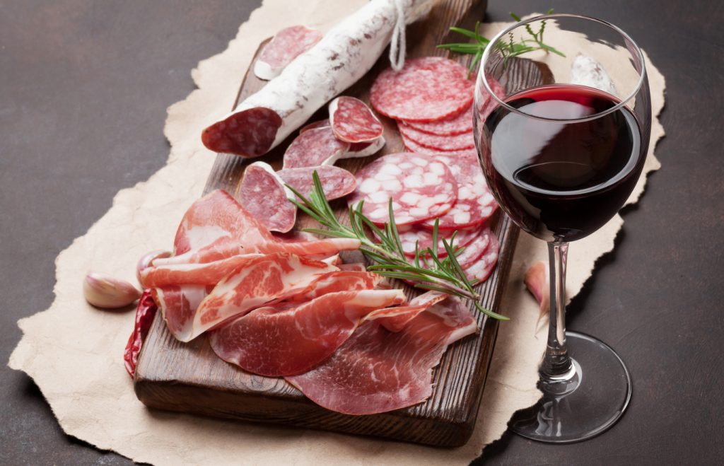 Salami, sliced ham, sausage, prosciutto, bacon and red wine glass. Meat antipasto platter on stone table